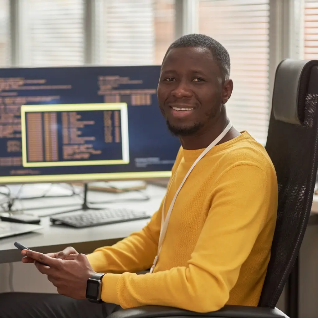 Man wearing a yellow sweater sitting in front of his office desk smiling at the camera.