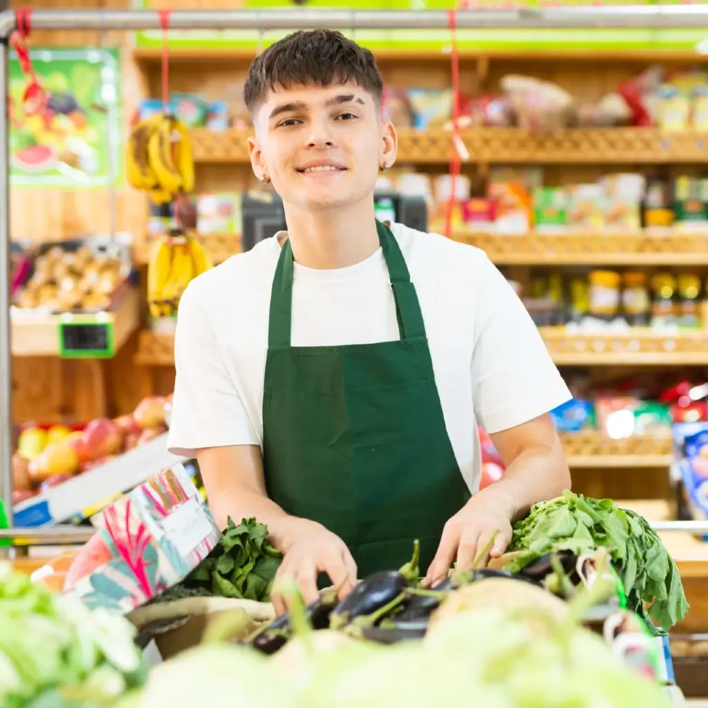 A young man in a white tee and apron arranging vegetable produce in a grocery.