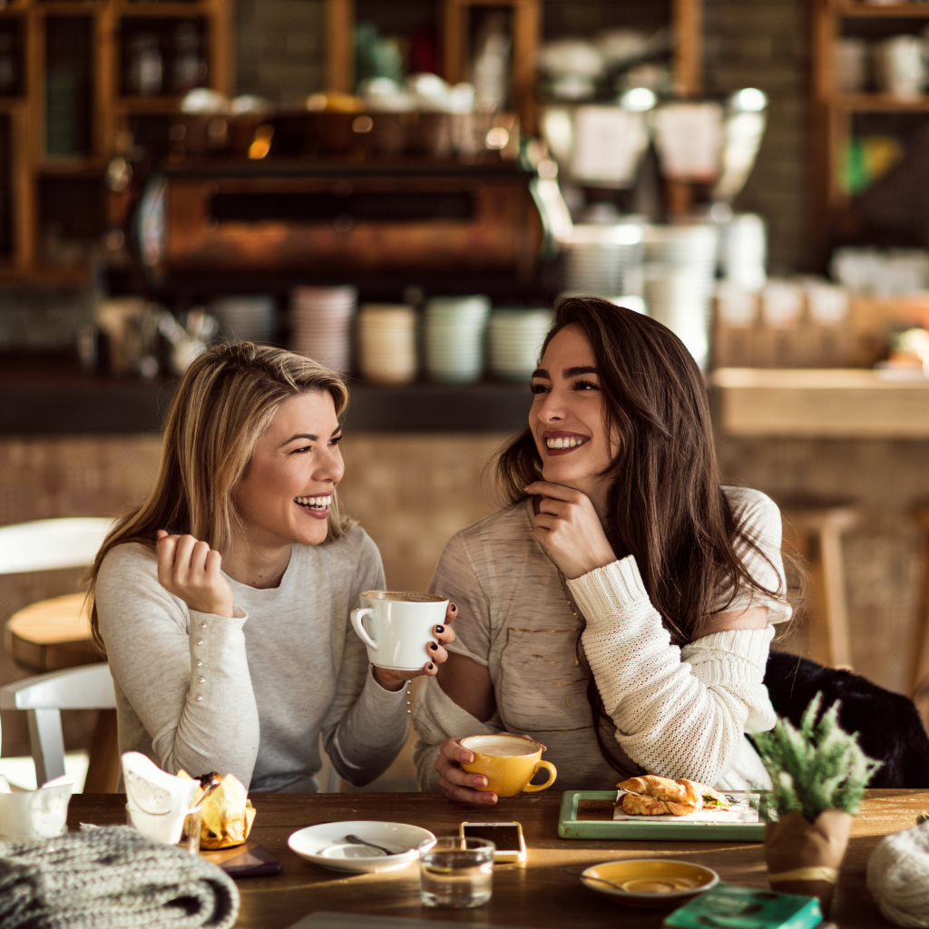 Two women laughing while having coffee and breakfast inside a cafe.