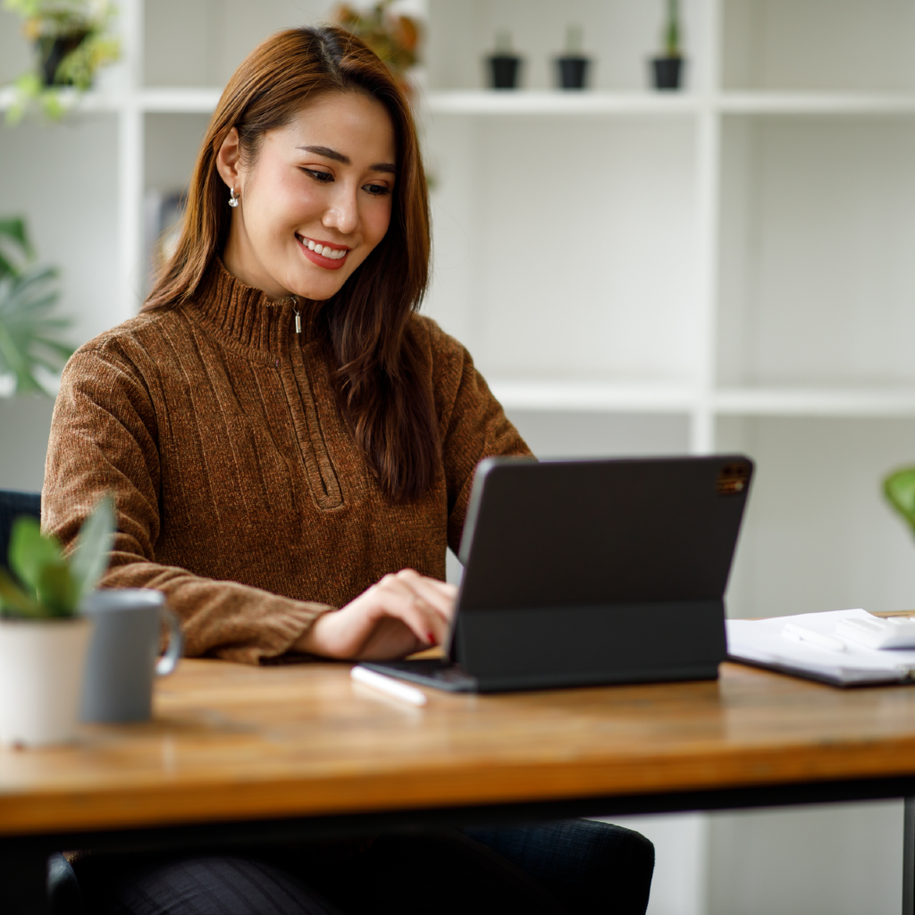 Woman smiling wide while typing on her tablet propped up on a table.