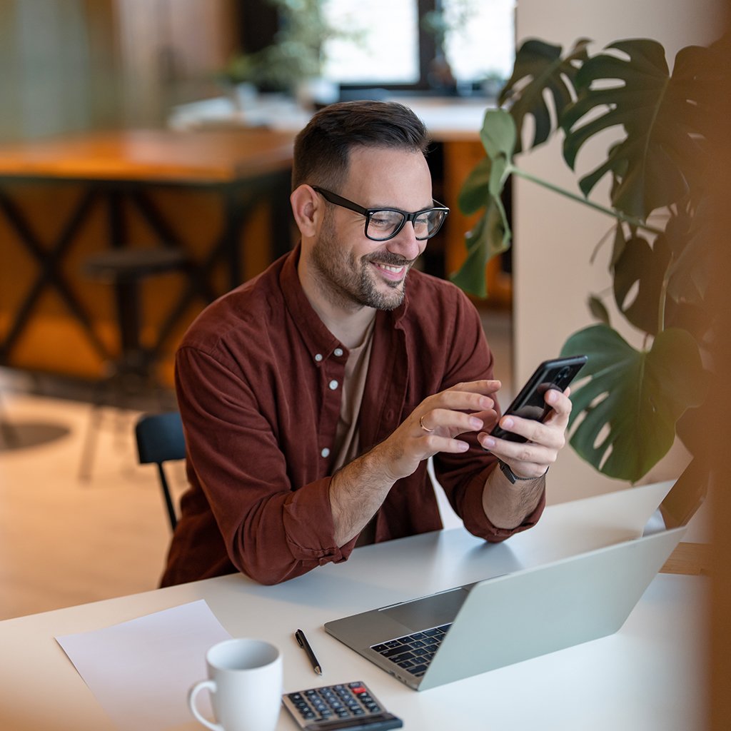 Man sitting in front of an open laptop while smiling and looking at his phone.