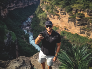 A man wearing sunglasses and a black shirt takes a selfie with a smartphone while standing near a canyon with a river below