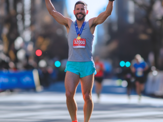 A male runner celebrates with arms raised after completing a marathon, wearing a medal and bib.