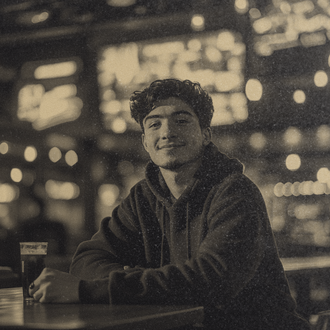 A young man in his late twenties sits at a high-top table in a lively sports bar, a smug grin on his lips. TV screens flicker with the Super Bowl game behind him.