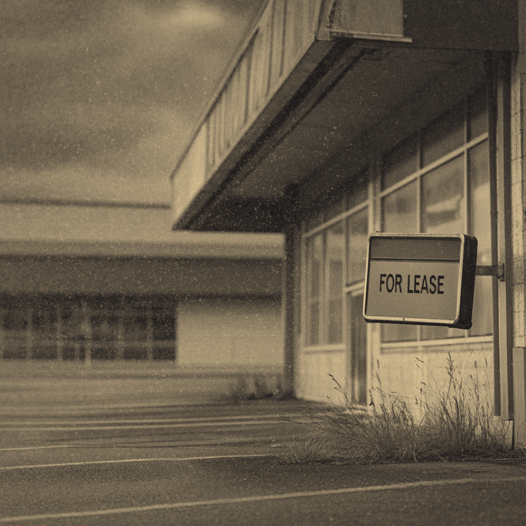 A deserted store with its signage removed, leaving faded outlines on the building. The empty parking lot is cracked, with weeds sprouting through the pavement. A "FOR LEASE" sign hangs in the front window.
